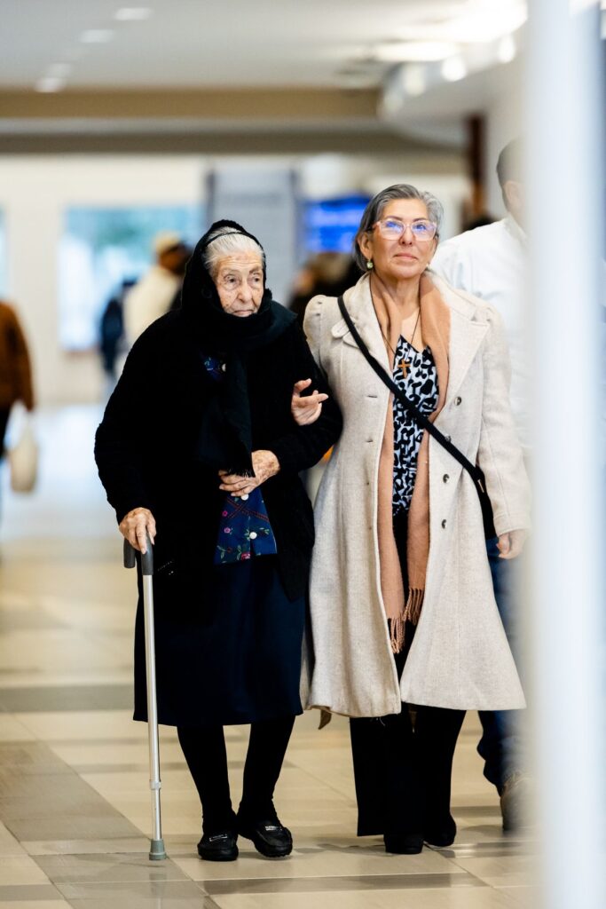 An older woman with a cane walking arm in arm with her daughter through a bright corridor