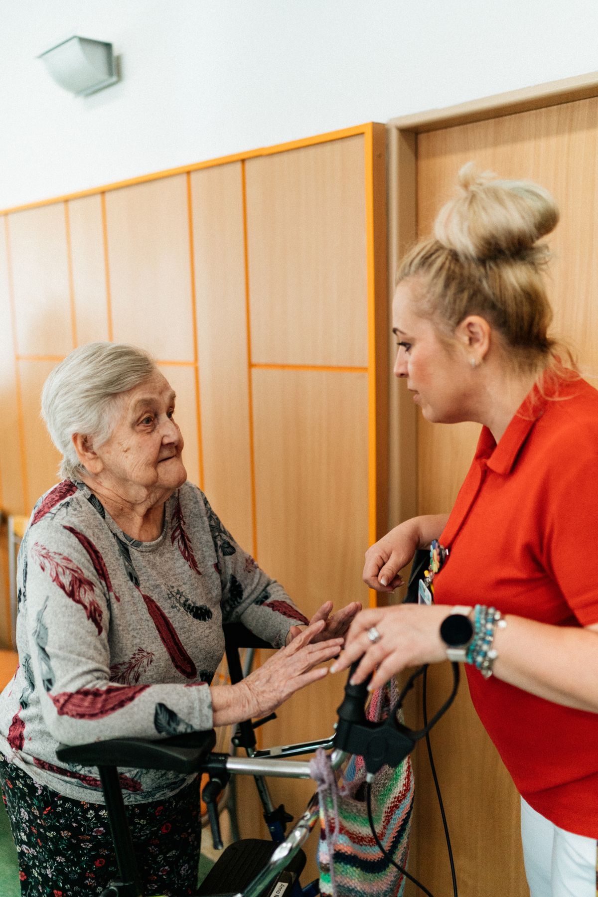 A caregiver in a red polo shirt leaning in to talk with a resident who uses a walker